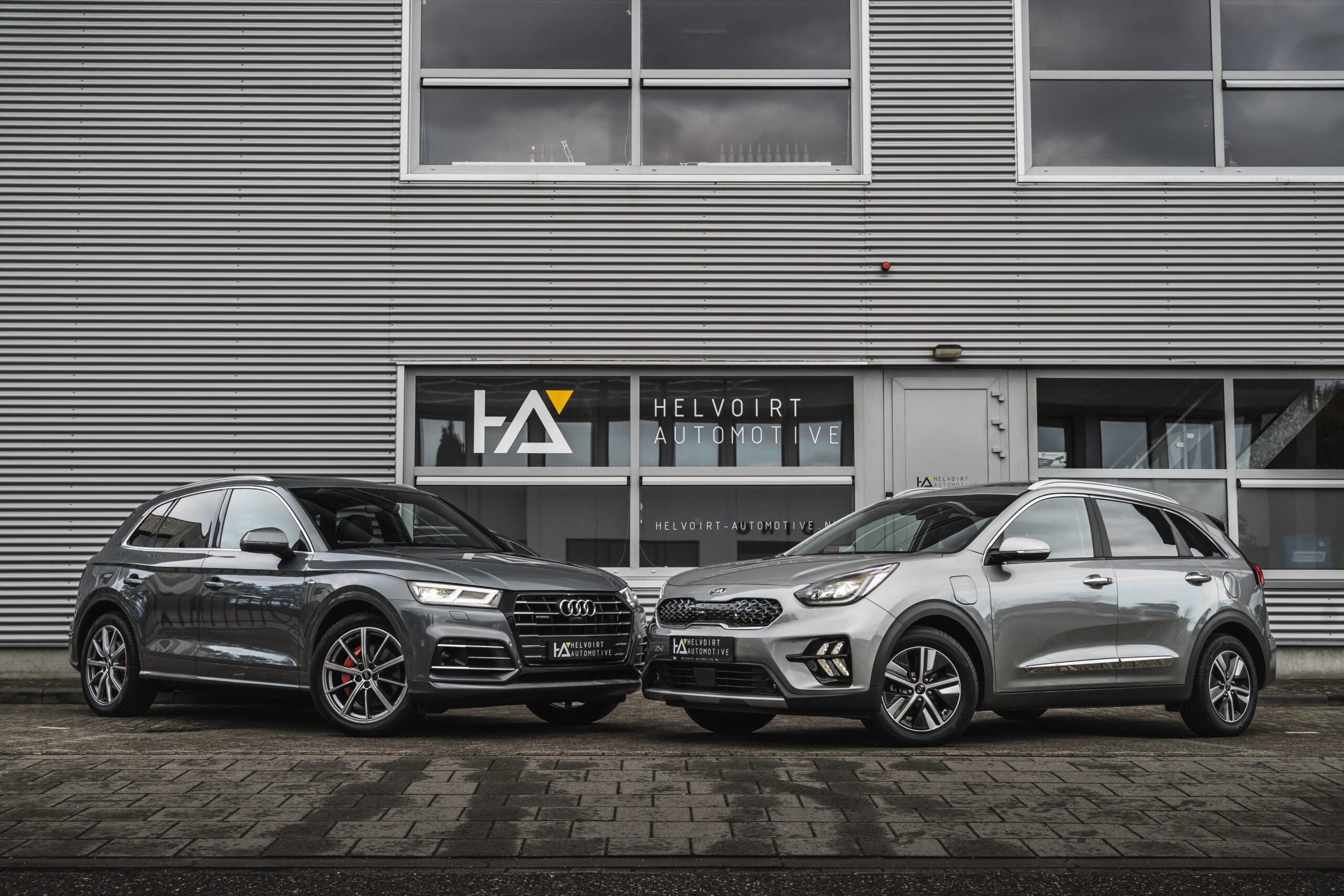 Two gray SUVs parked outside the Helvoirt Automotive showroom on a wet street.